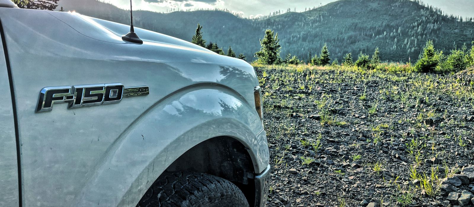 A white pickup parked at the edge of a cleared cut, forested ridges and cloud beyond