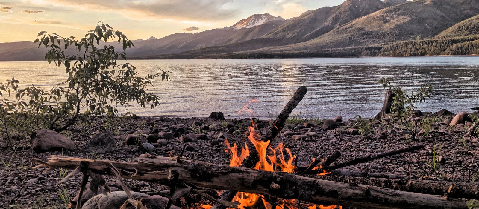 A small fire burning on a driftwood gravel bar at dusk, a lake and blue mountain ranges behind