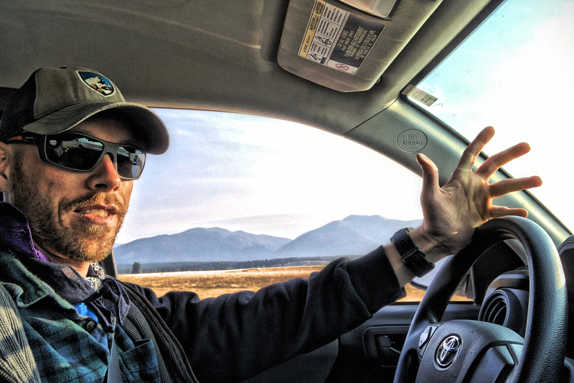 Mark Ulett driving a truck through a Montana valley, one hand off the wheel mid-sentence, mountains and cured grass through the windshield